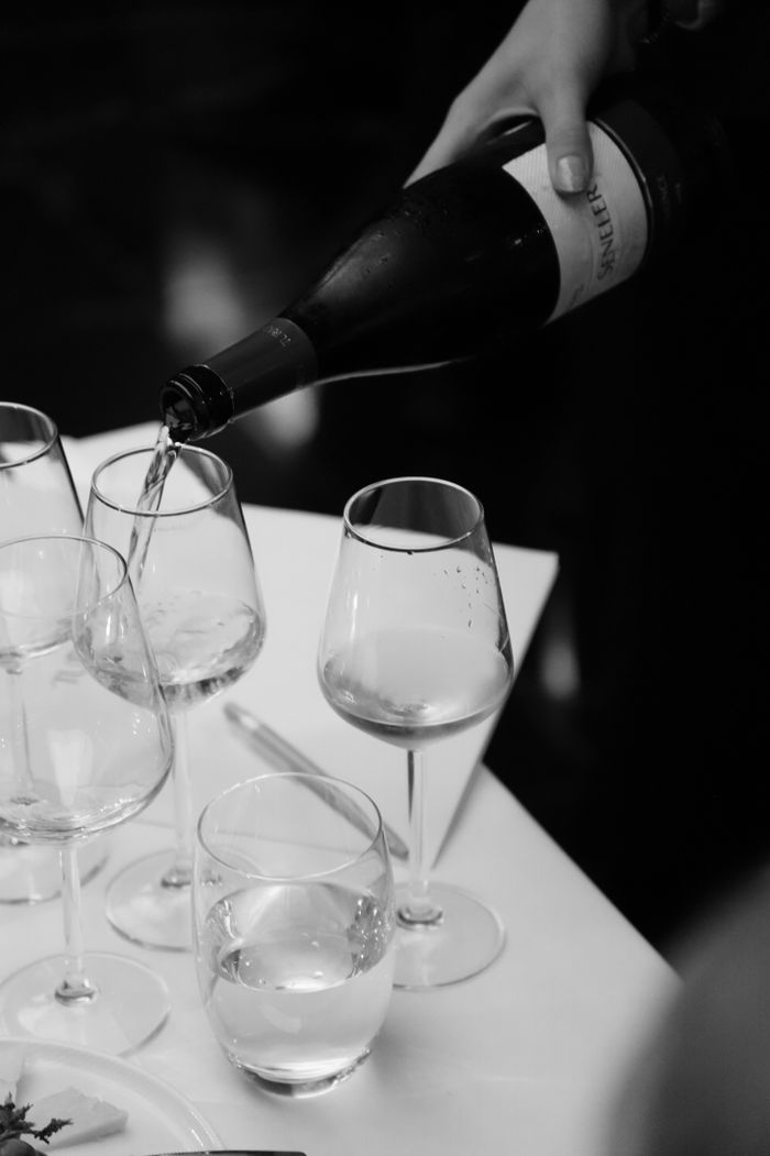 A monochrome image of wine being elegantly poured into glasses at a dining table.